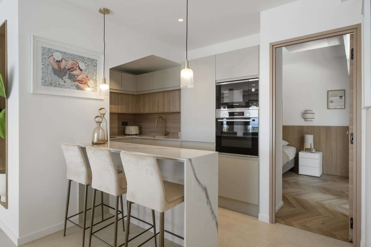 A bright, modern kitchen features a marble island with bar stools, integrated appliances, and wood accents, opening to a bedroom with a wooden floor.