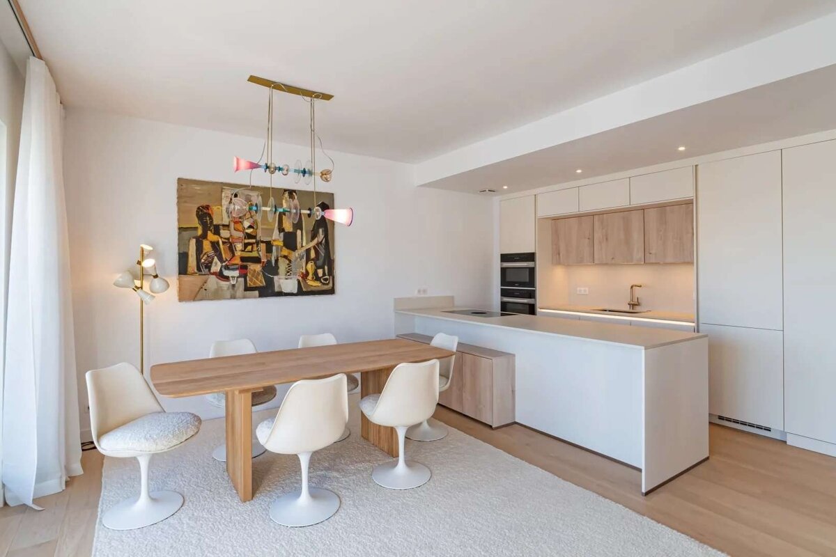 Modern open-plan dining room and kitchen. Features a wooden table, white tulip chairs, abstract art, and a colorful chandelier over sleek white/wood cabinetry.