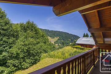 A sunny balcony view of a lush green mountain vista, with dense trees, grassy hills, and distant chalets under a clear blue sky.
