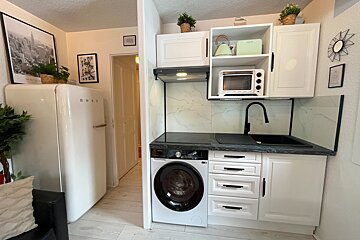 A compact, modern white kitchen features a Smeg fridge, washing machine, stovetop, sink, microwave, and kettle, with black countertops and wall art.