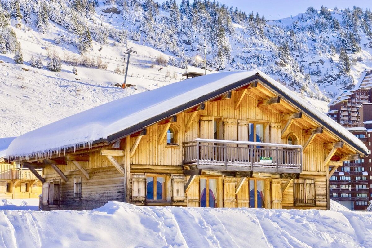 A wooden chalet covered in snow, set against a backdrop of snowy alpine mountains and pine trees.
