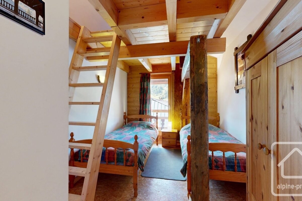 A cozy, rustic wooden bedroom features two single beds, exposed beams, a ladder to a loft, and a window with an outdoor view.