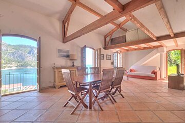 A spacious room with exposed wooden beams, terracotta floor, dining table, sofa area, and large arched doors opening to a scenic bay and mountains.