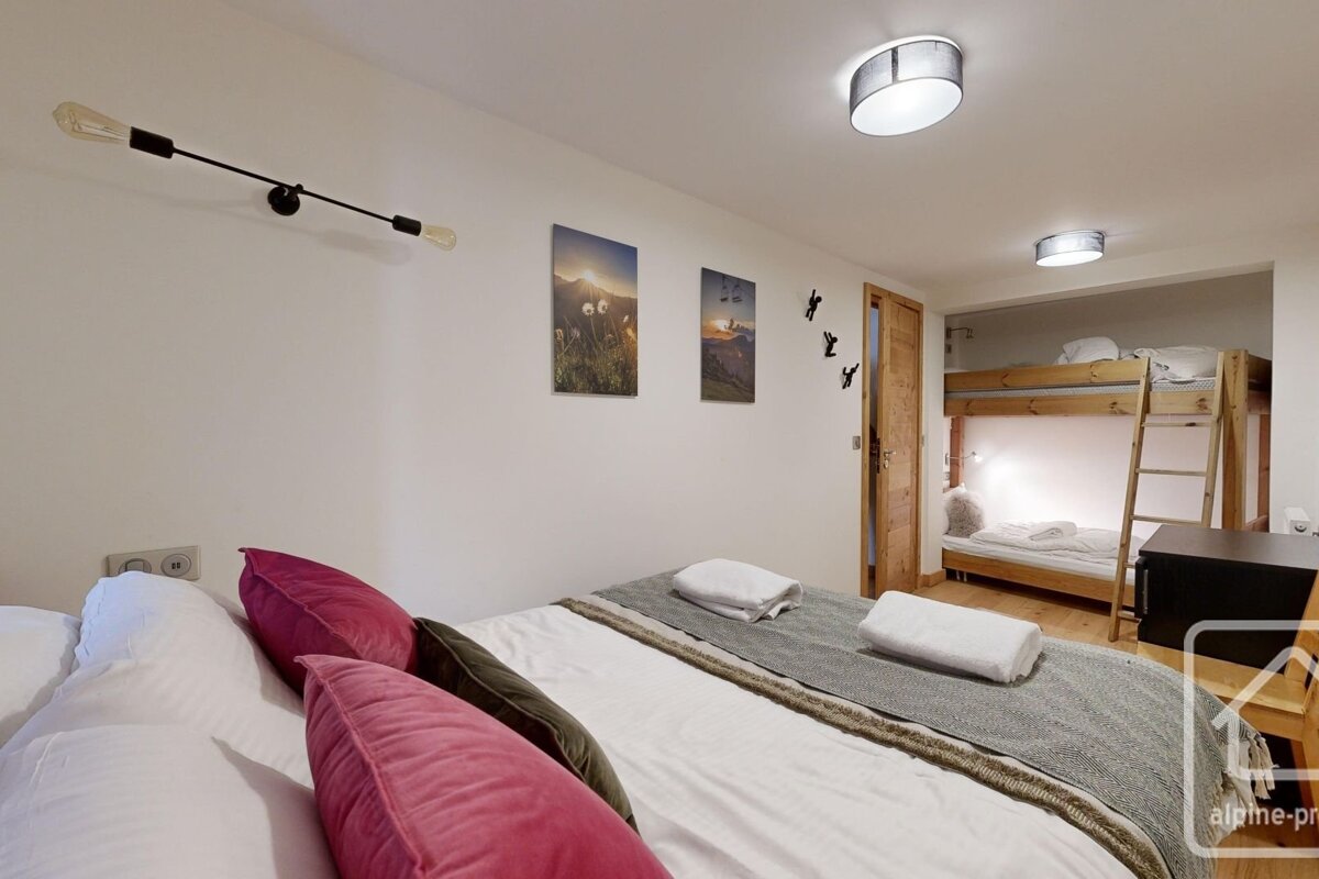 A modern bedroom features a large bed with pink pillows, mountain wall art, and wooden bunk beds in an alcove. White walls and warm lighting create a cozy space.