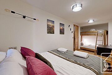 A modern bedroom with a main bed, pink pillows, and bunk beds in an alcove. White walls feature mountain art and skier hooks.