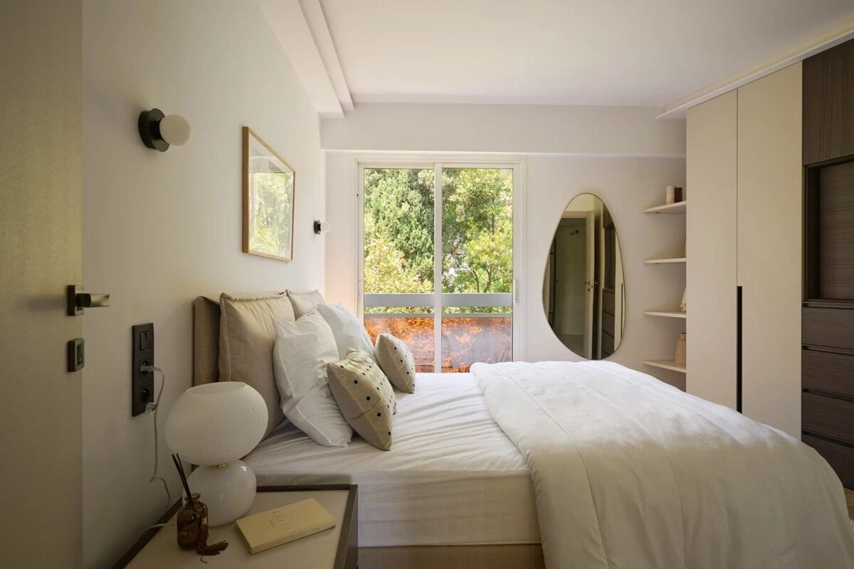 Modern minimalist bedroom in neutral tones, featuring a white bed, an oval wall mirror, and a large window overlooking lush green trees.