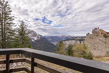Scenic view of mountains, pine trees, and a cliffside resort under a cloudy sky, seen from a wooden balcony.