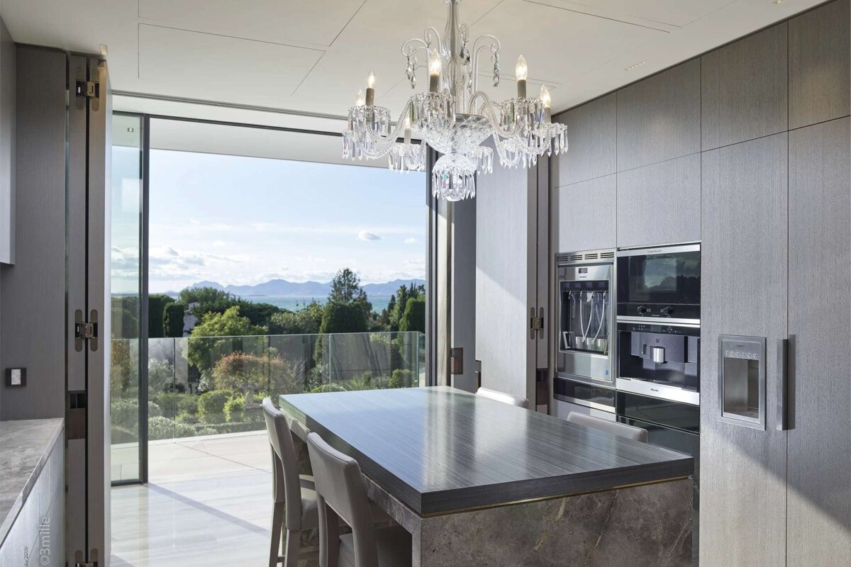 Luxurious modern kitchen with a crystal chandelier, island, and large windows opening to a stunning ocean and mountain view.