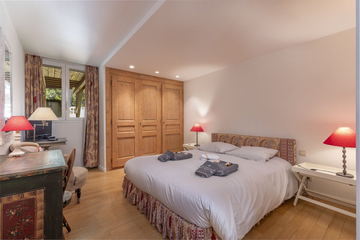 A cozy, rustic bedroom featuring a large double bed, wooden closets, red bedside lamps, and an antique desk.