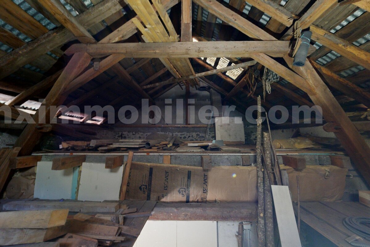 A cluttered attic space with exposed wooden roof beams, visible roof structure, and insulation rolls among various stored items. A watermark is present.