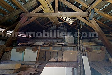 A cluttered attic space with exposed wooden roof beams, visible roof structure, and insulation rolls among various stored items. A watermark is present.