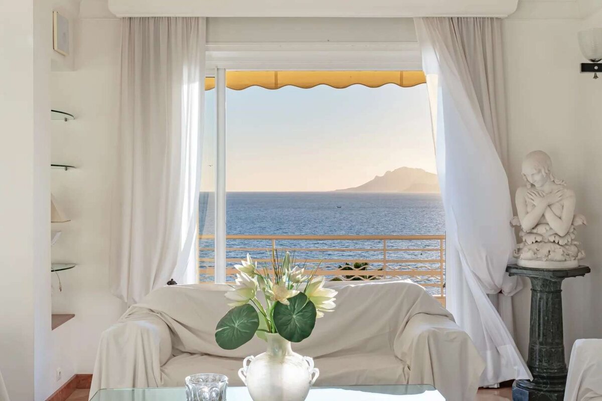 A bright room with white decor and a sofa opens to a balcony with a stunning sea view and distant mountains under a warm sky.