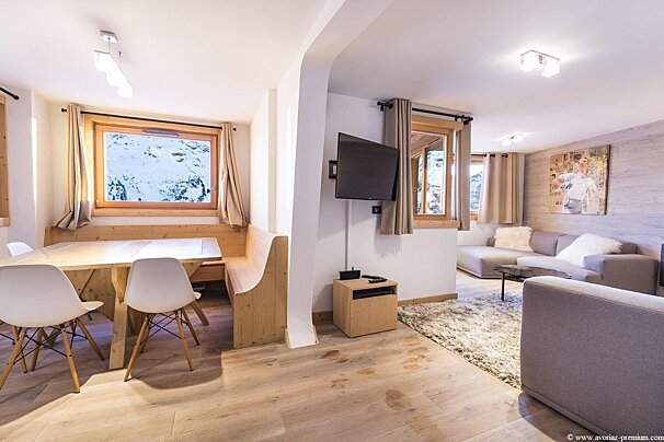 Modern cabin living/dining area with light wood floors. Dining nook overlooks snowy mountains; living room features gray sofas.