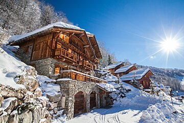Snow-covered wooden chalets on a sunny mountain slope under a bright blue sky, creating a picturesque winter scene.