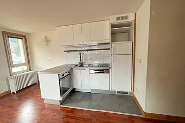 Bright, unfurnished room featuring a compact white kitchenette, warm wood floor, and a window with a large radiator.