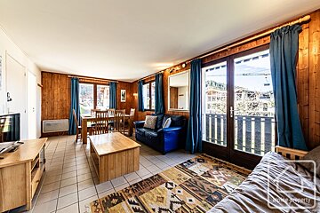 A rustic living/dining area with wood paneling, blue curtains, tiled floor, wooden furniture, a blue sofa, and a patterned rug, offering outdoor views.