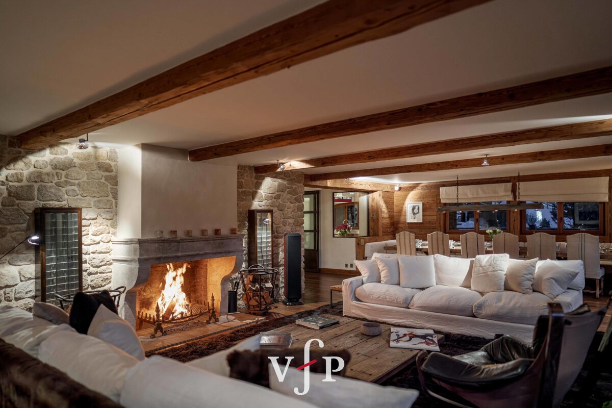 Cozy chalet living room featuring a warm stone fireplace, exposed wooden ceiling beams, and comfortable white plush sofas.