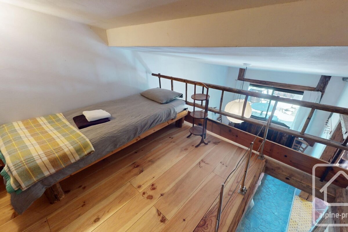 A cozy loft bedroom features a single bed with a plaid blanket on a wooden floor, overlooking a lower level through a rustic wooden railing.