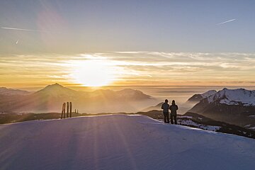 Two people standing on top of a snowy mountain at sunset