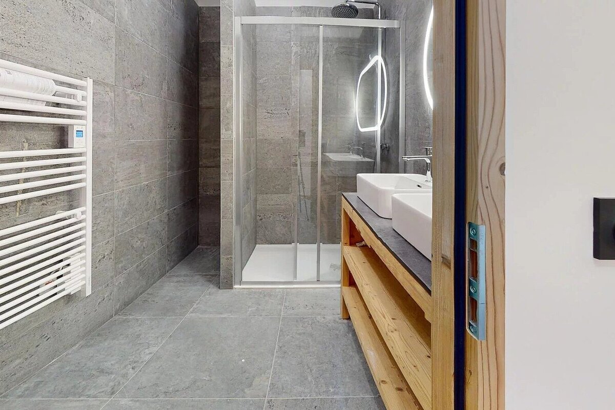A modern bathroom with grey tiled walls and floor features a glass shower, double sinks on a wooden vanity, backlit mirrors, and a towel warmer.