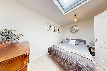 A bright bedroom with a double bed, wooden dresser, and skylight. White walls are decorated with framed art and a round mirror.