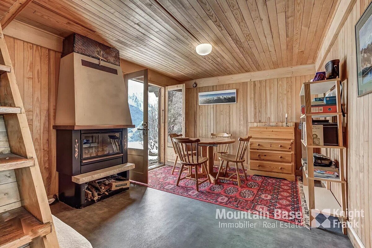 A cozy, wood-paneled room with a fireplace, dining area, and a view of snowy mountains through an open door. Features a red rug and rustic wooden furniture.