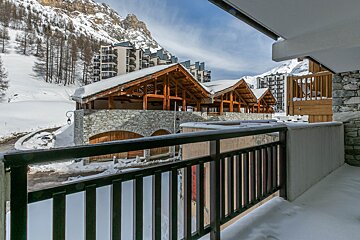 A scenic winter view from a balcony overlooking a snow-covered alpine village with charming stone/wood buildings, modern apartments, and distant mountains.