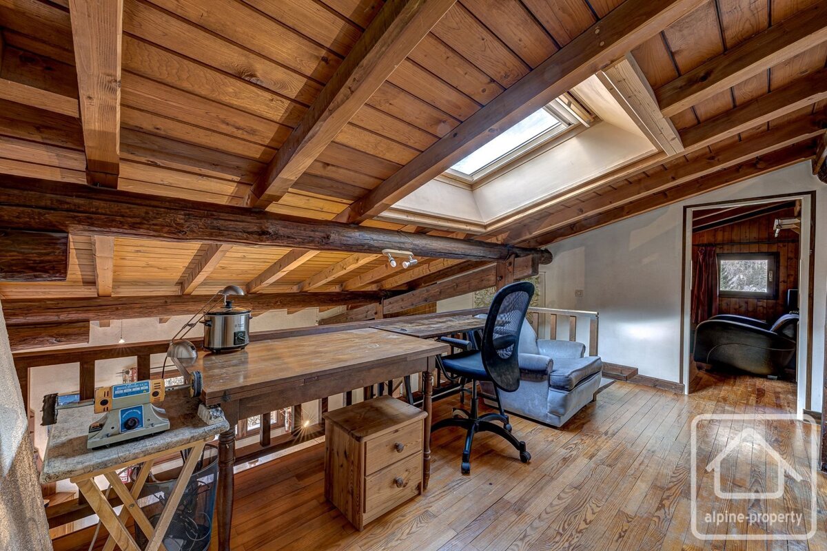 An attic home office with wood-planked ceilings and beams, featuring a desk, office chair, armchair, and skylight window.