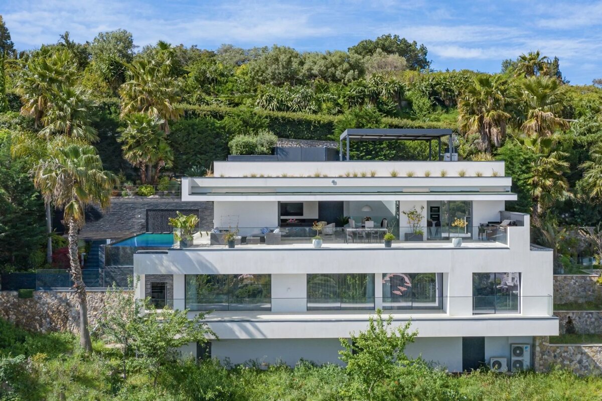 Modern white multi-level villa with a swimming pool, nestled on a lush green hillside with palm trees.