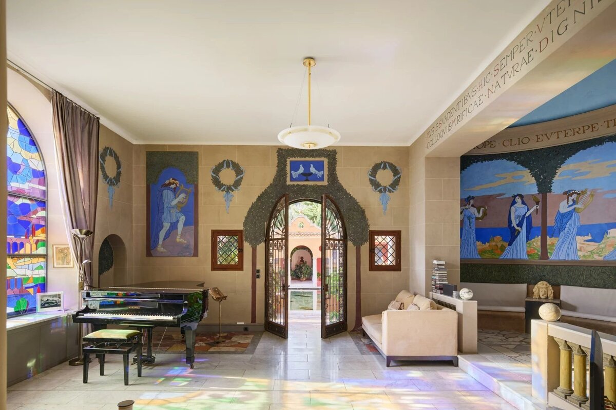 Elegant interior featuring classical murals, a grand piano by a stained-glass window, and arched doors opening to a sunlit courtyard.