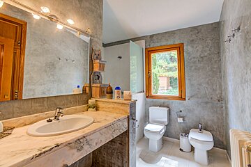 A bathroom with grey textured walls, a marble vanity, a large mirror, a wooden-framed window, and a toilet with a bidet.