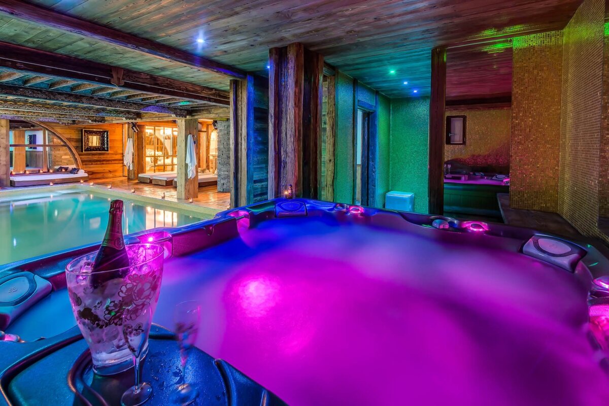 Luxurious indoor spa featuring a vibrant purple-lit hot tub with champagne, alongside a swimming pool under rustic wooden beams.