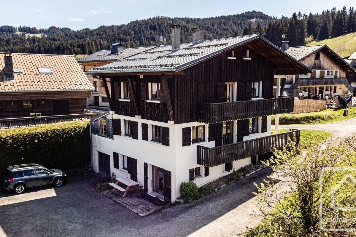 A white and dark wooden chalet-style house with balconies and shutters sits in a sunny, forested mountain landscape with other houses and a parked car.