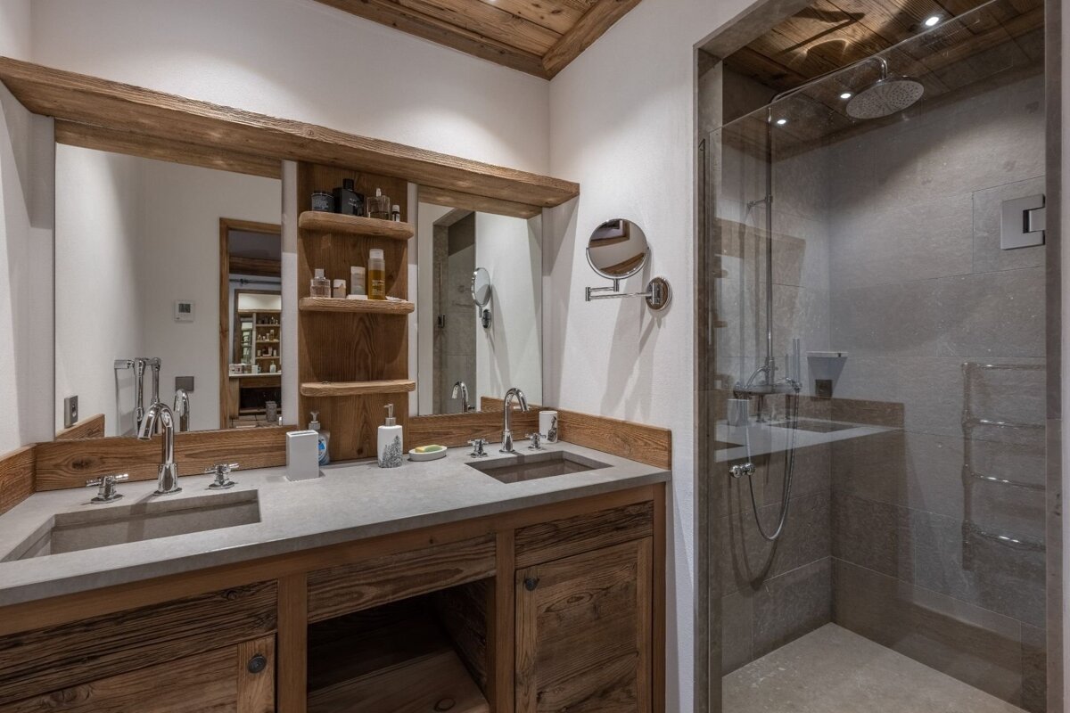 A rustic modern bathroom with a double wood vanity, stone sinks, large mirror, and a glass-enclosed shower. Wooden ceiling adds warmth.