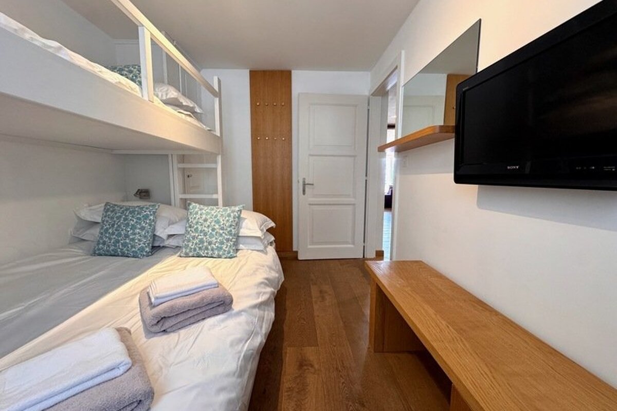 Clean bedroom with white bunk beds, patterned pillows, hardwood floors, a wall-mounted TV, and a long wooden bench.