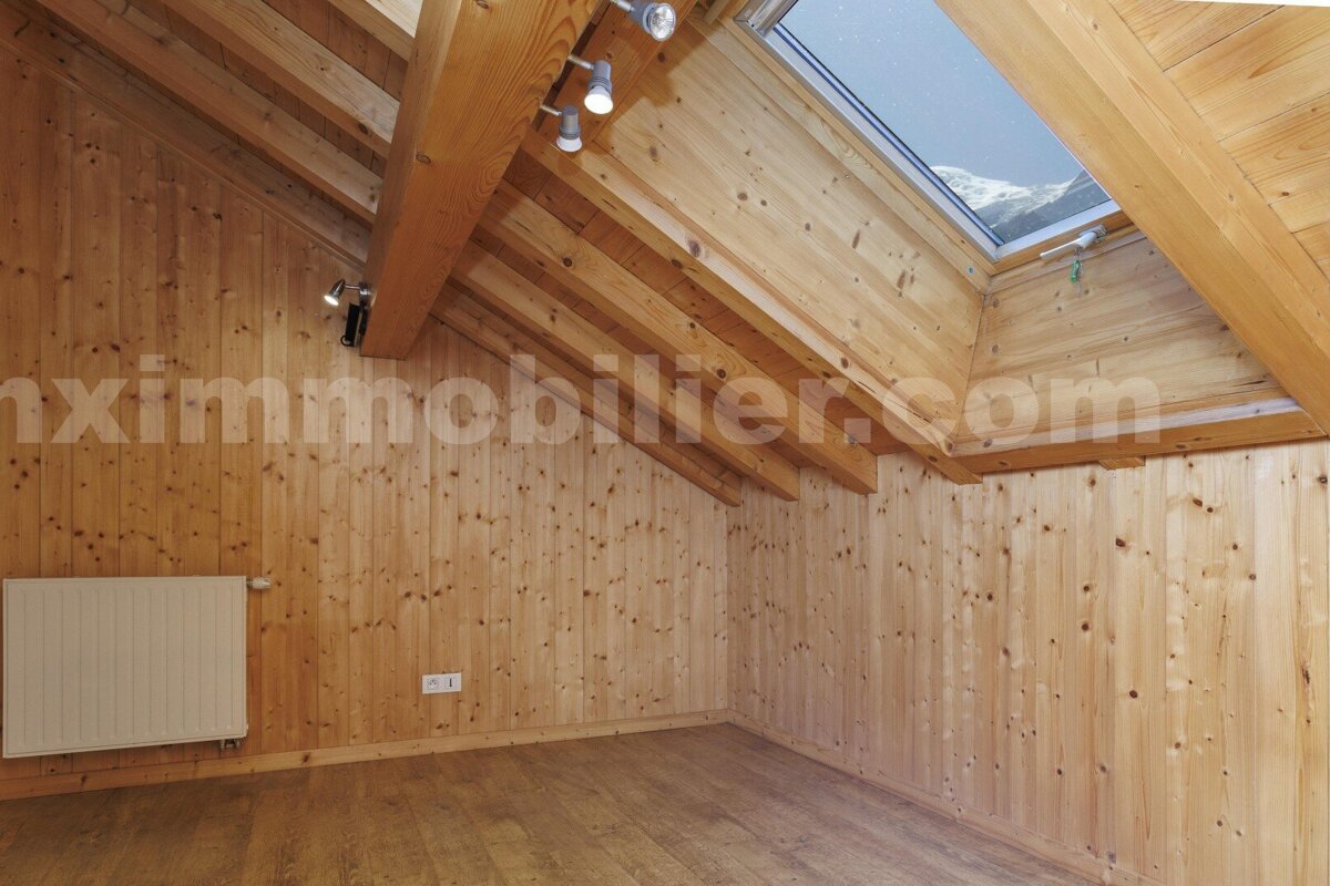 A cozy attic room with light wood paneling, exposed beams, and a wooden floor. A skylight reveals a view of snowy mountains. Includes a radiator and spotlights.