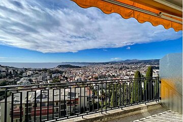 A vibrant view from a balcony showing a coastal city, the sea, and mountains under a blue sky with white clouds, framed by an orange awning.