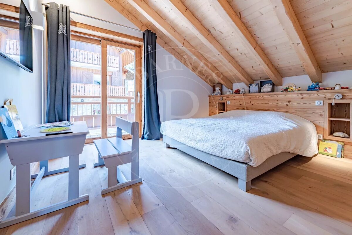 A rustic-chic bedroom features a large bed, wooden beamed ceiling, and a child's desk. A balcony offers views of other wooden buildings.