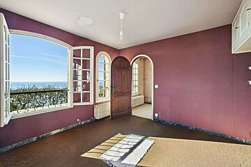 An empty room with plum-colored walls, stunning ocean views through open windows, an arched wooden door, and a ceiling water stain.