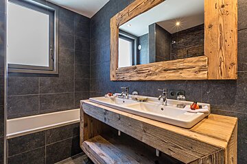 Stylish bathroom with dark tiled walls, a rustic wooden double vanity & mirror frame, and a white bathtub.