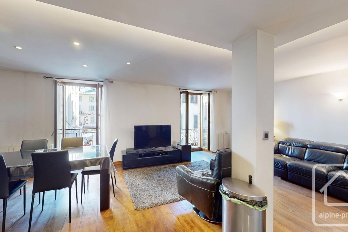A bright living/dining room with hardwood floors, white walls, a dining table, TV, black leather seating, and balcony access.