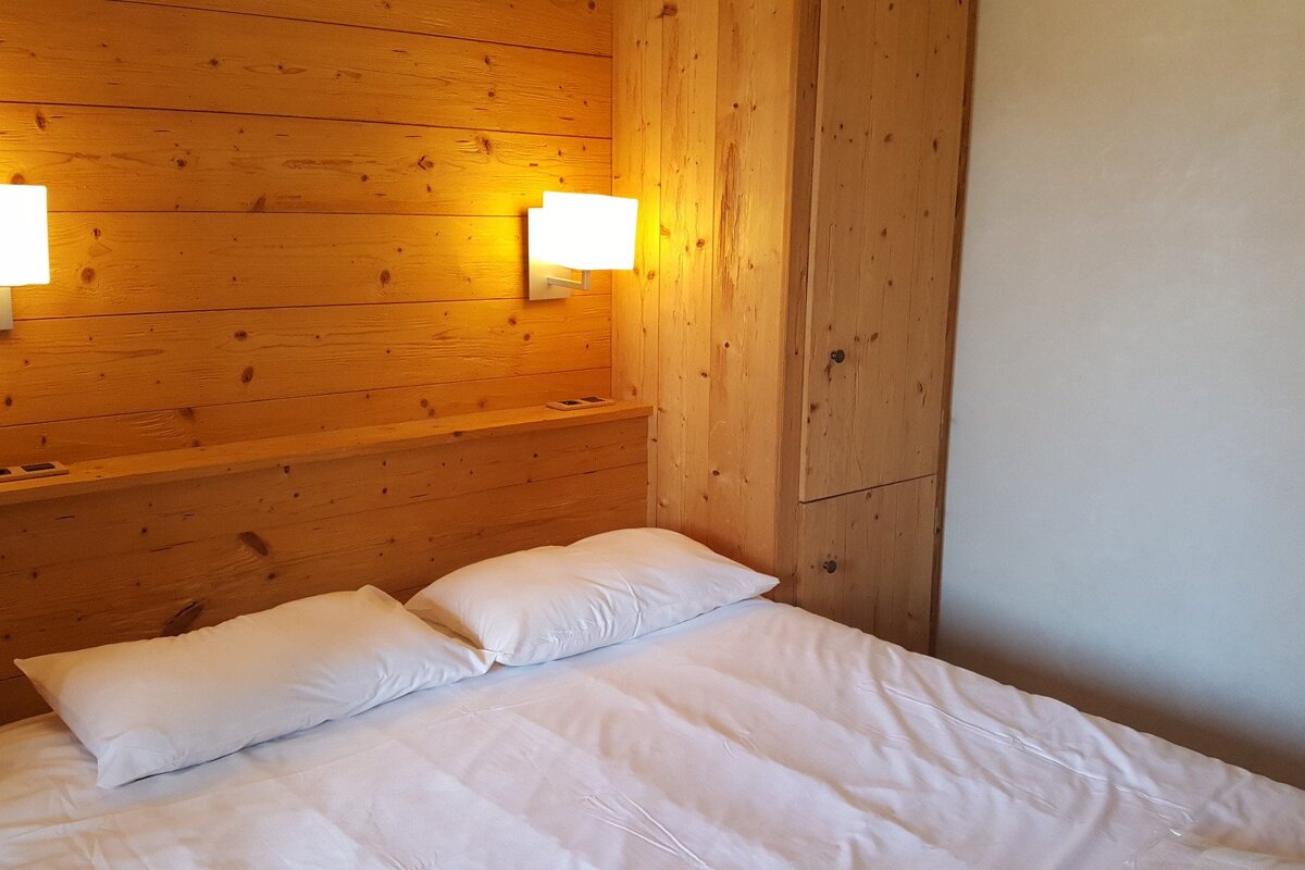 A cozy bedroom featuring a bed with white linens and pillows against a warm wooden headboard and paneled wall, with an integrated wooden cabinet.