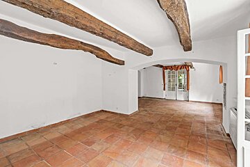 Empty room with terracotta floor, white walls, and exposed wooden beams. An archway leads to French doors with orange curtains.