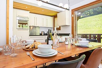 A set wooden dining table in a modern kitchen, next to a large window overlooking a lush green hillside.