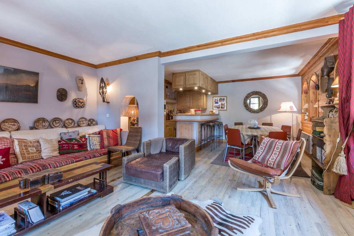 A rustic, open-plan living and dining area with ethnic decor, light wood floors, a cozy sofa, and a kitchen bar in the background.