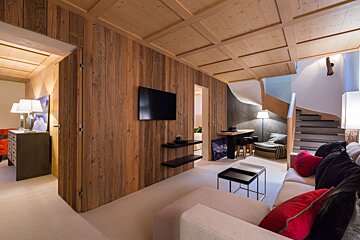 A cozy, chalet-style living room with extensive wood paneling, a modern sofa, wall-mounted TV, dining nook, and a sleek spiral staircase.
