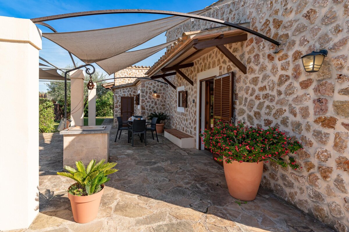 A stone house with a shaded patio, featuring an outdoor dining area, potted plants, and a bright blue sky overhead.