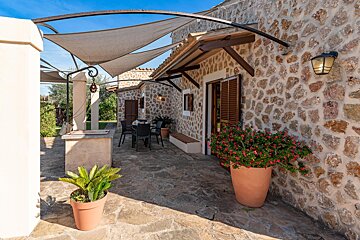 A stone house with a shaded patio, featuring an outdoor dining area, potted plants, and a bright blue sky overhead.