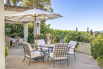 An inviting sunny patio features wrought iron furniture, a round table with an umbrella, and abundant green foliage under a clear blue sky.