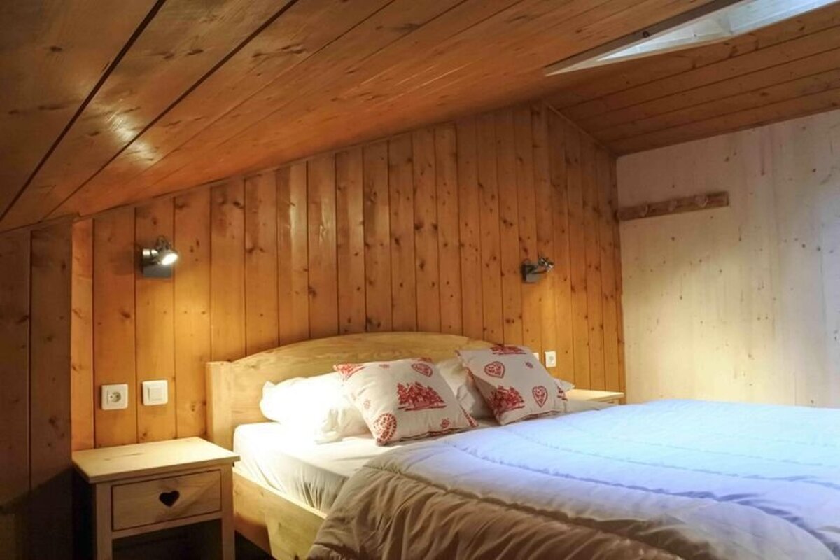 A cozy, rustic bedroom featuring warm wood-paneled walls and a slanted ceiling. A bed with patterned pillows and bedside lamps creates an inviting ambiance.